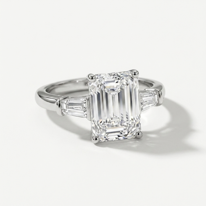 An emerald-cut diamond ring with baguette side stones in a white gold metal setting, photographed on a clean white background with a soft shadow.