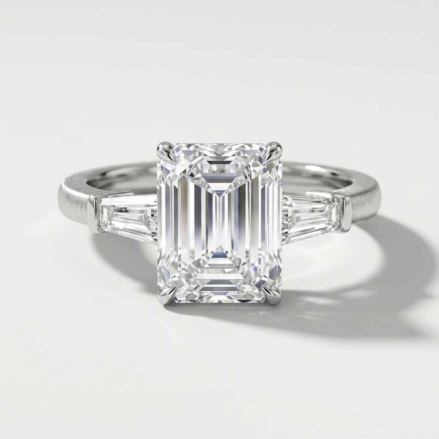 An emerald-cut diamond ring with baguette side stones in a white gold setting, photographed on a clean light gray background with a soft shadow.