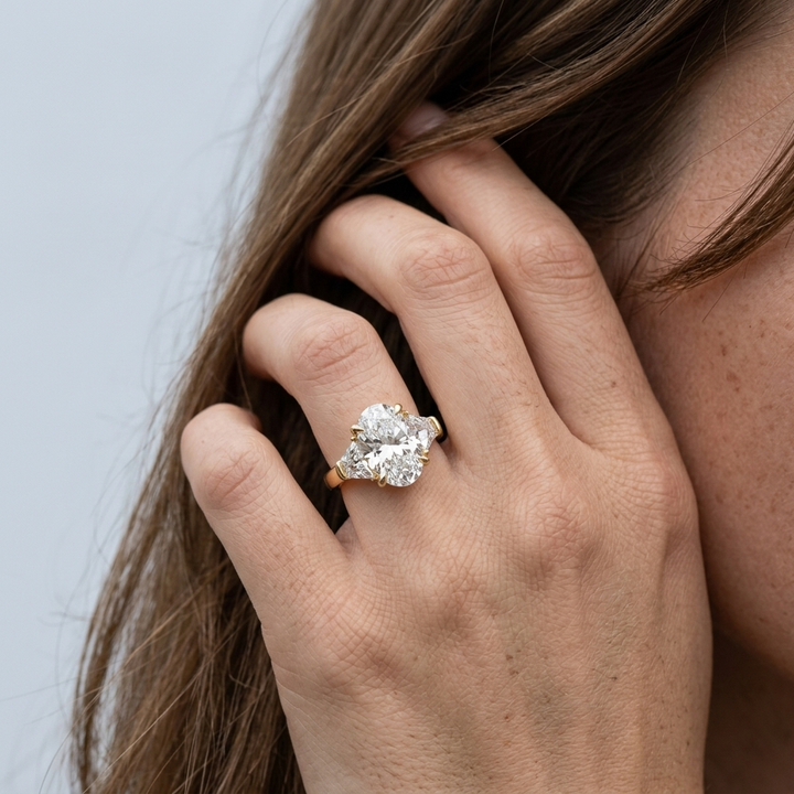 A close-up of a woman's hand wearing a large Oval diamond engagement ring with a yellow gold band against a soft gray background.