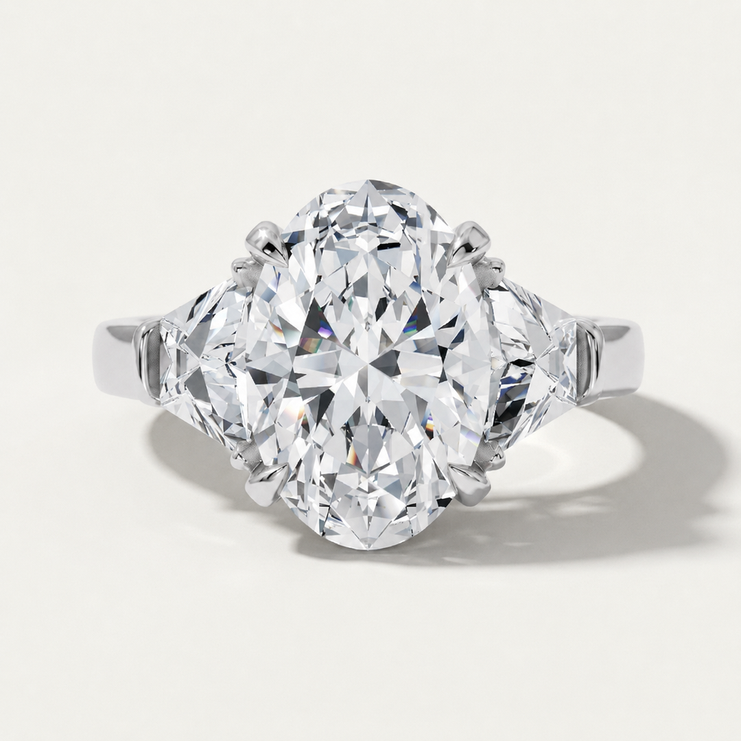 An oval-cut diamond ring with a brilliant center stone and two smaller diamond-shaped side stones, set in a polished white gold metal band, shown on a light off-white background with a soft shadow.