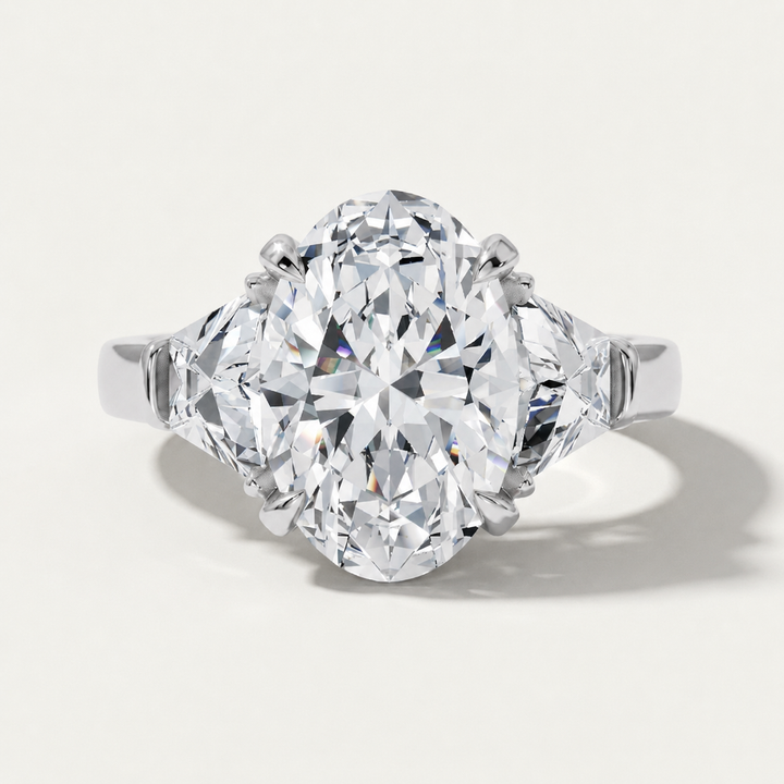 An oval-cut diamond ring with a brilliant center stone and two smaller diamond-shaped side stones, set in a polished white gold metal band, shown on a light off-white background with a soft shadow.