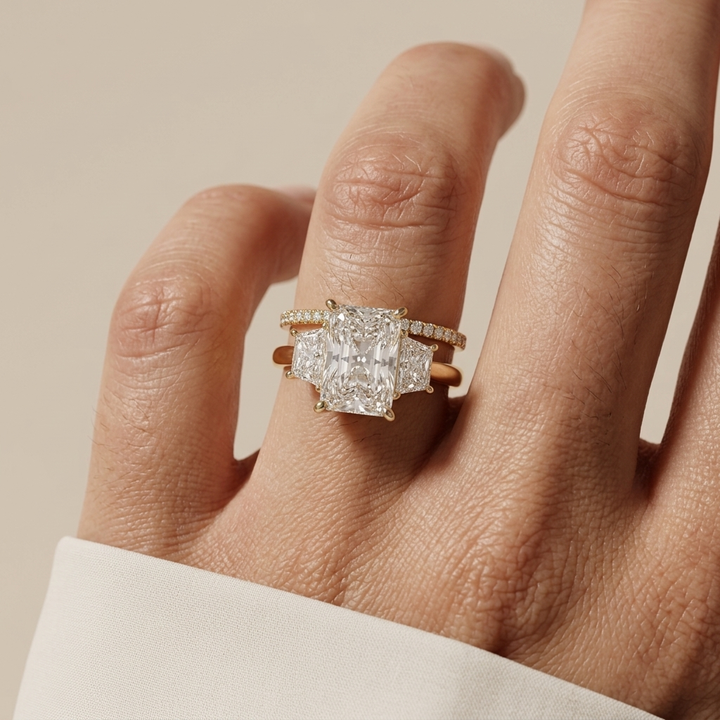 A close-up of a hand wearing a yellow gold ring set with three Radiant-cut diamonds and smaller side diamonds, against a soft beige background.