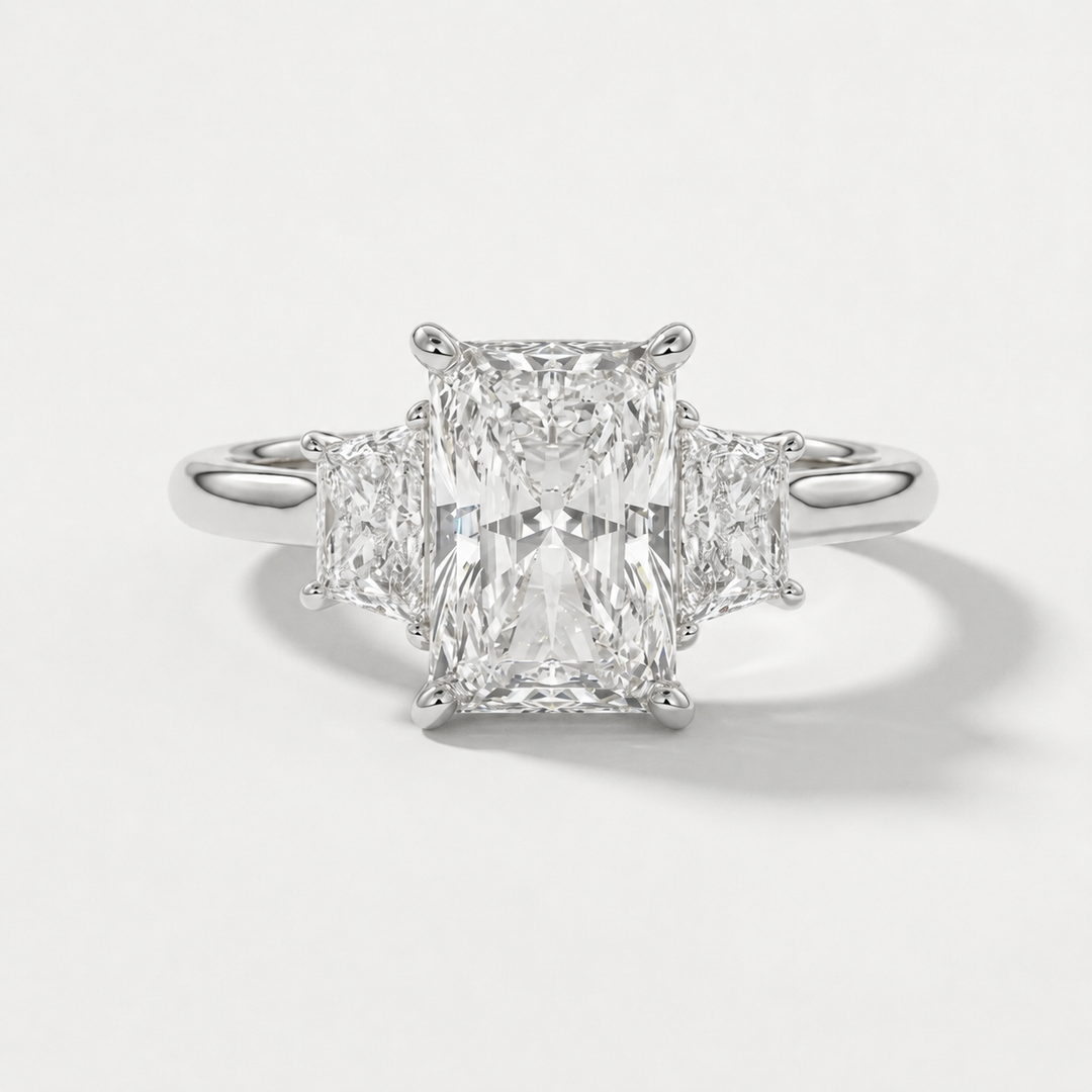 A radiant-cut three-stone diamond ring with a brilliant center stone and two smaller side stones set in a polished white gold metal band, shown on a light gray background.