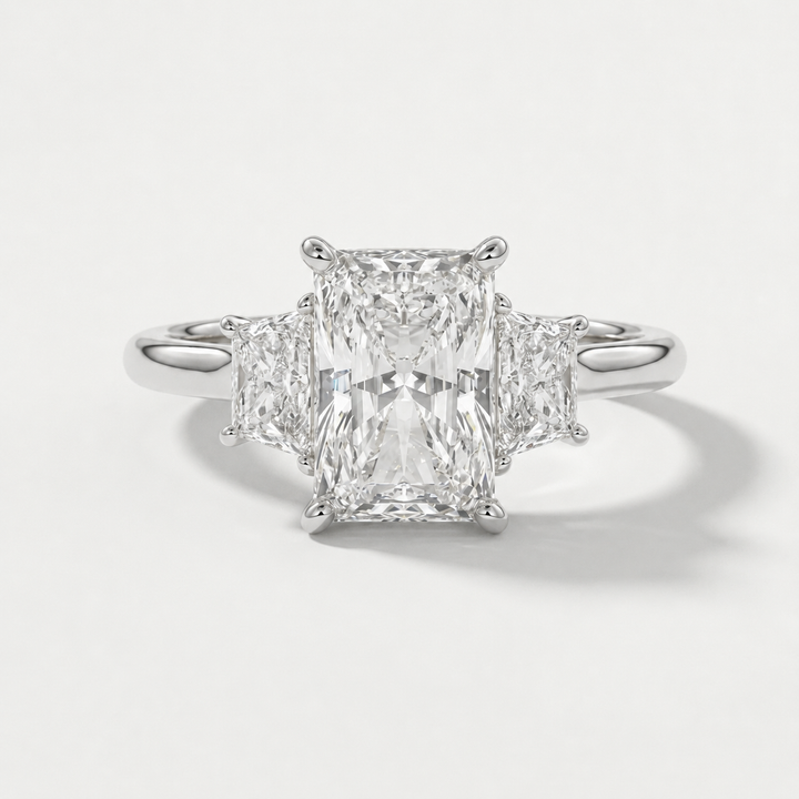A radiant-cut three-stone diamond ring with a brilliant center stone and two smaller side stones set in a polished white gold metal band, shown on a light gray background.