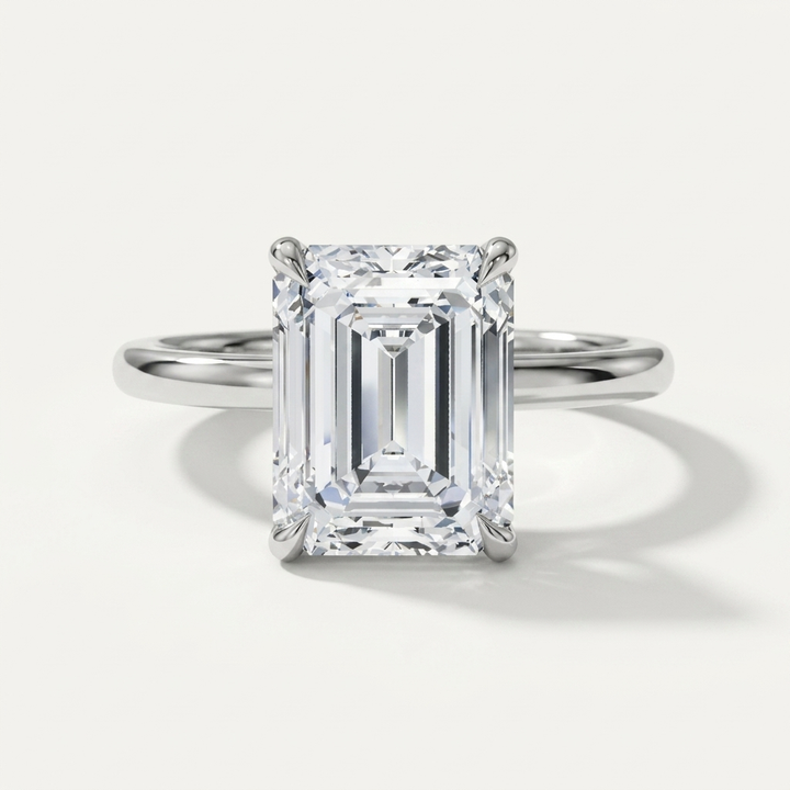An emerald-cut diamond solitaire ring in a polished white gold metal setting, centered on a clean, light off-white background with a soft shadow.