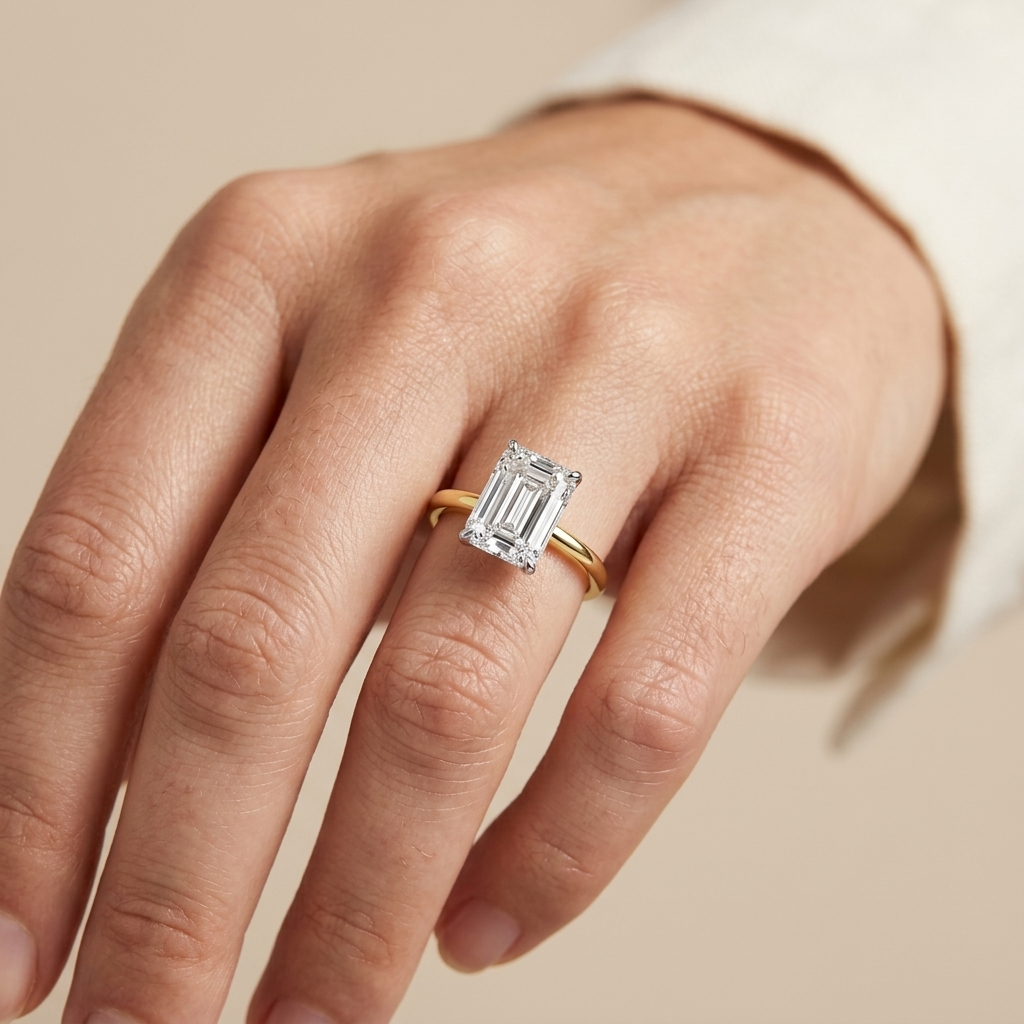 A close-up of a hand wearing a yellow gold ring with an emerald-cut diamond center stone, set against a soft beige background.