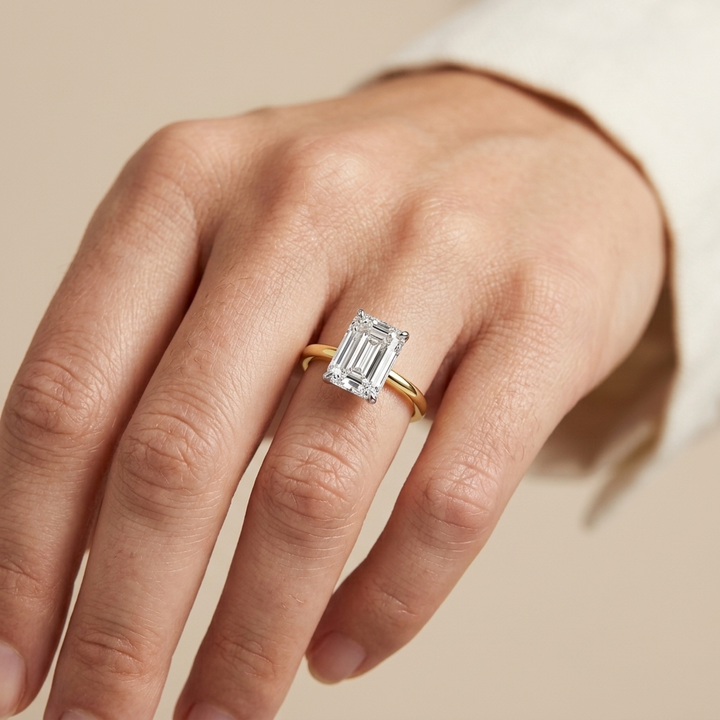 A close-up of a hand wearing a yellow gold ring with an emerald-cut diamond center stone, set against a soft beige background.