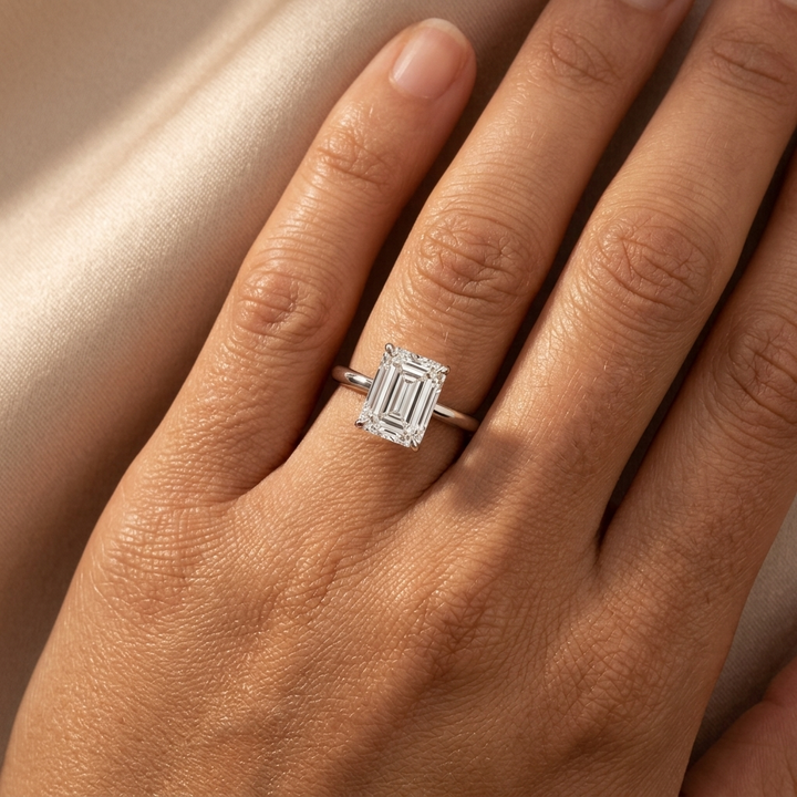 A close-up of a hand wearing a silver engagement ring with a rectangular baguette-cut diamond. The ring is set on a soft, light beige satin fabric background.