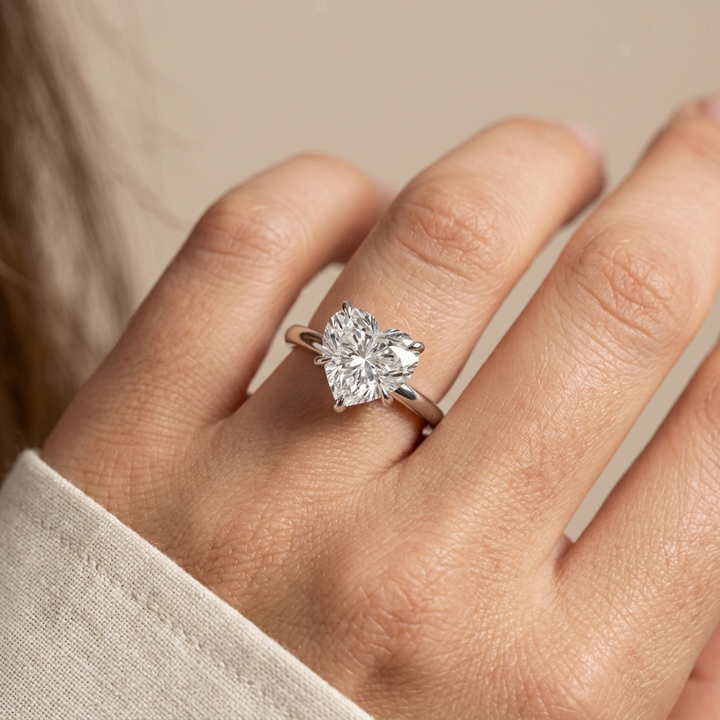 A heart-shaped diamond engagement ring on a white gold band, shown worn on a person's ring finger against a soft beige background.