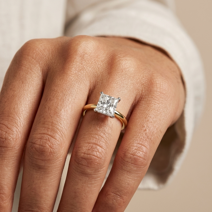 A close-up of a hand wearing a gold ring with a princess cut diamond, set against a soft beige background.