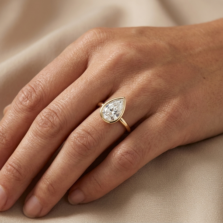 A close-up of a hand wearing a gold ring with a pear-cut diamond, resting on a soft beige fabric background.