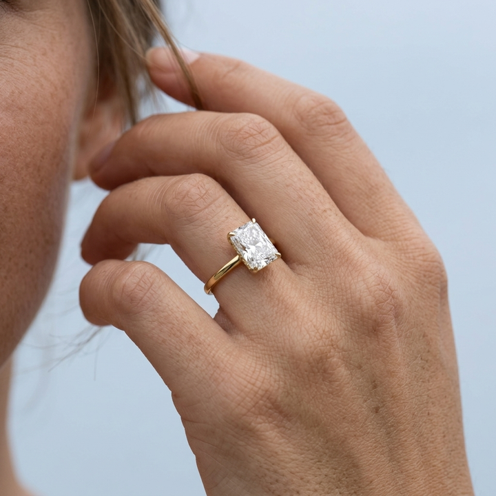 A close-up of a person's hand wearing a yellow gold ring with a rectangular radiant diamond, set against a soft light blue background.