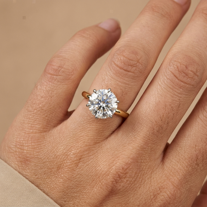 A close-up of a hand wearing a yellow gold ring with a round brilliant-cut diamond. The ring is shown on the ring finger against a soft beige background.