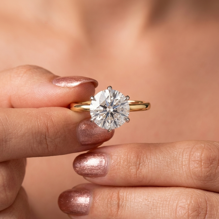 A close-up of a hand with metallic rose gold nail polish holding a classic solitaire engagement ring with a round brilliant-cut diamond and a yellow gold band.