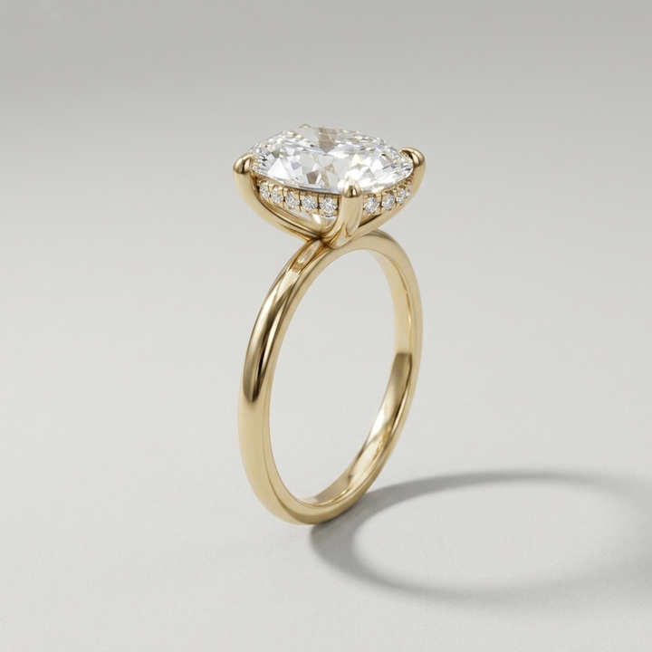 A yellow gold engagement ring featuring a large oval diamond center stone in a prong setting, with small round stones along the hidden halo, shown on a neutral light gray background.