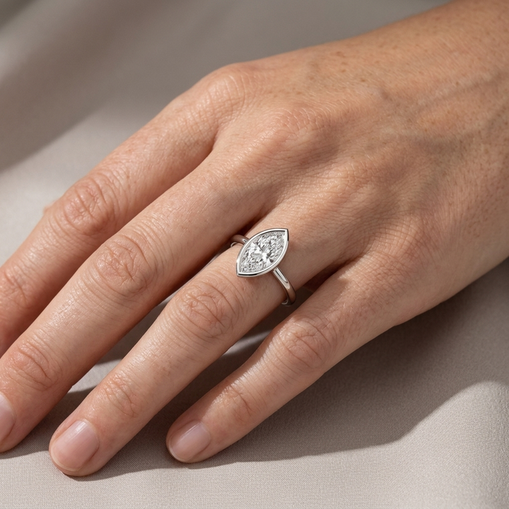 A close-up of a hand wearing a white gold marquise-shaped diamond ring, resting on a neutral beige fabric surface.