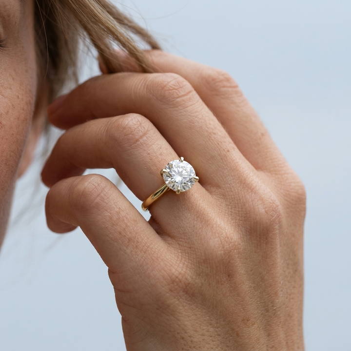 A close-up of a person's hand wearing a classic solitaire engagement ring with a round brilliant-cut diamond set in a yellow gold band against a soft, light blue-gray background.