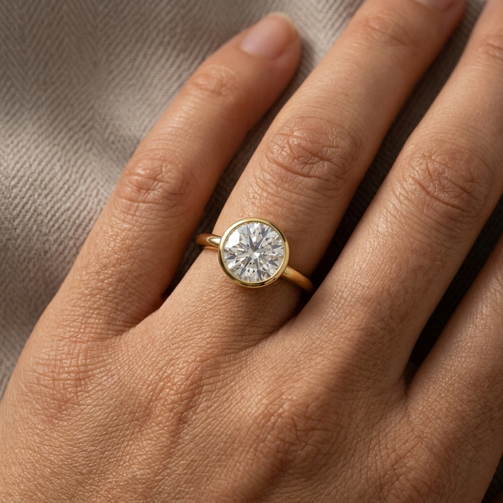 A close-up of a gold ring with a round brilliant-cut bezel set diamond set on a person's finger, resting against a textured beige fabric background.