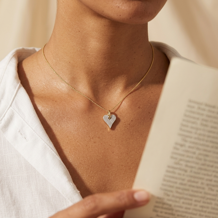 A close-up lifestyle shot of a model wearing a delicate gold chain necklace with a small, pave-set heart-shaped pendant. The model is wearing a white linen top and holding an open book, with warm, soft lighting highlighting the jewelry.